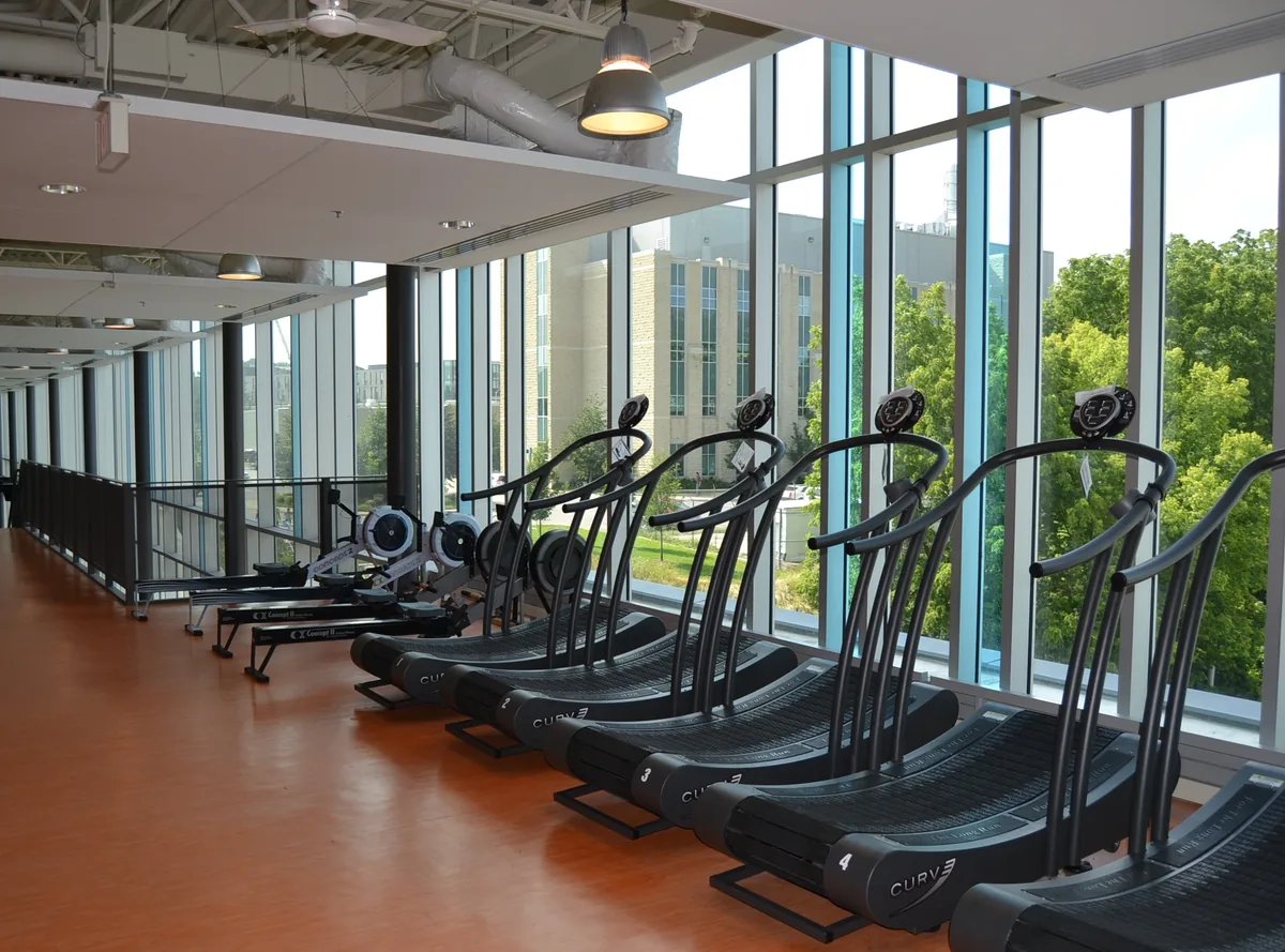 Salle de fitness lumineuse avec tapis de course et rameurs devant de grandes baies vitrées.